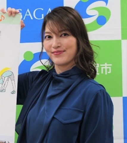 青と緑の市松模様に佐賀市のロゴが並ぶ背景の前で、光沢のある紺色のブラウスを着たロングヘアの朝夏まなとさんが、ストレッチのイラストが描かれた白いパネルを右手に持ちながら、正面を見つめている上半身の写真