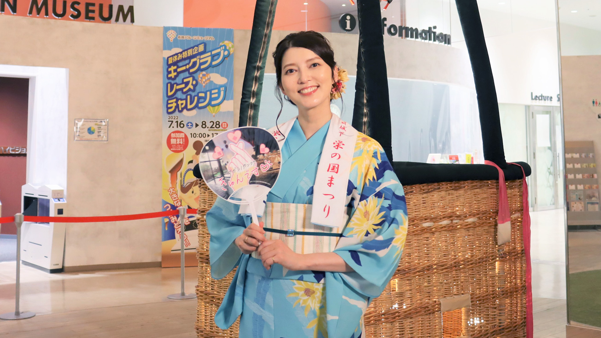熱気球のバスケットの横で、水色に黄色い花柄の浴衣を着た朝夏まなとさんが「佐賀城下栄の国まつり」のタスキをかけ、うちわを手にして微笑む、バルーンミュージアムの館内を背景にした記念写真