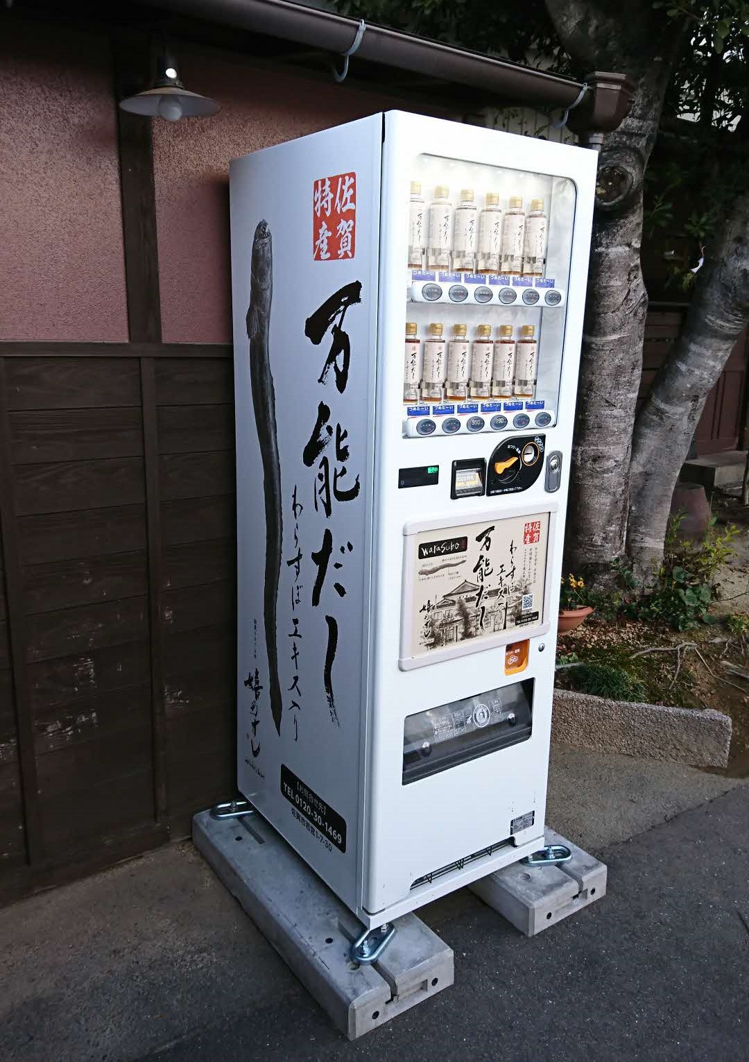 屋外の木の幹や建物の壁際に設置された、佐賀特産「わらすぼエキス入り 万能だし」の大きな魚のイラストと商品名が側面に描かれた白い自販機が、コンクリートの土台の上に固定されている様子を捉えた写真