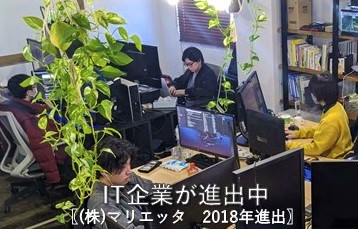 「IT企業が進出中【株式会社マリエッタ 2018年進出】」の文字と、観葉植物の緑が映える明るいオフィス内で、数人の社員がデスクに向かいパソコン作業に集中している様子の写真