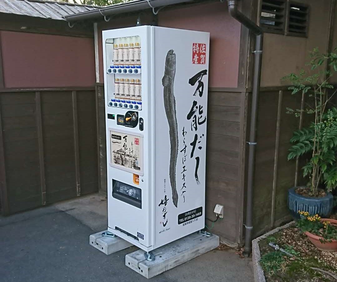 和風の建物の壁際に設置された、佐賀特産「わらすぼエキス入り 万能だし」の大きな魚のイラストと商品名が側面に描かれた白い自販機が、コンクリートの土台の上に固定され、周囲に植栽や鉢植えが並ぶ様子を捉えた写真