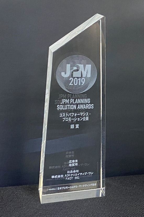 「JPM 2019」のロゴと共に「JPM PLANNING SOLUTION AWARDS 銀賞」や「佐賀市」、「株式会社 ADKクリエイティブ・ワン」等の刻印が入った、透明なクリスタルトロフィーの写真