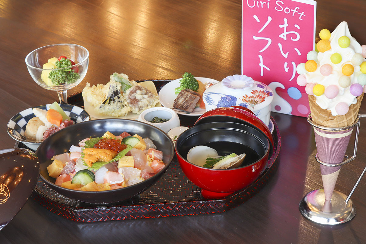 お盆の上に海鮮丼、蛤のお吸い物、天ぷら、煮物などの豪華な和食膳が並び、その傍らにはカラフルな伝統菓子おいりがトッピングされたソフトクリームが添えられた、贅沢で色彩豊かな料理を写した写真