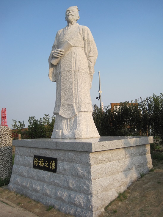 澄み渡る青空を背景に、右手に巻物を持ちゆったりとした中国の伝統的な衣装を纏って佇む、石造りの「徐福之像」が立派な石組みの台座の上に設置され、周囲には緑の木々が並ぶ屋外の静かな風景を捉えた写真