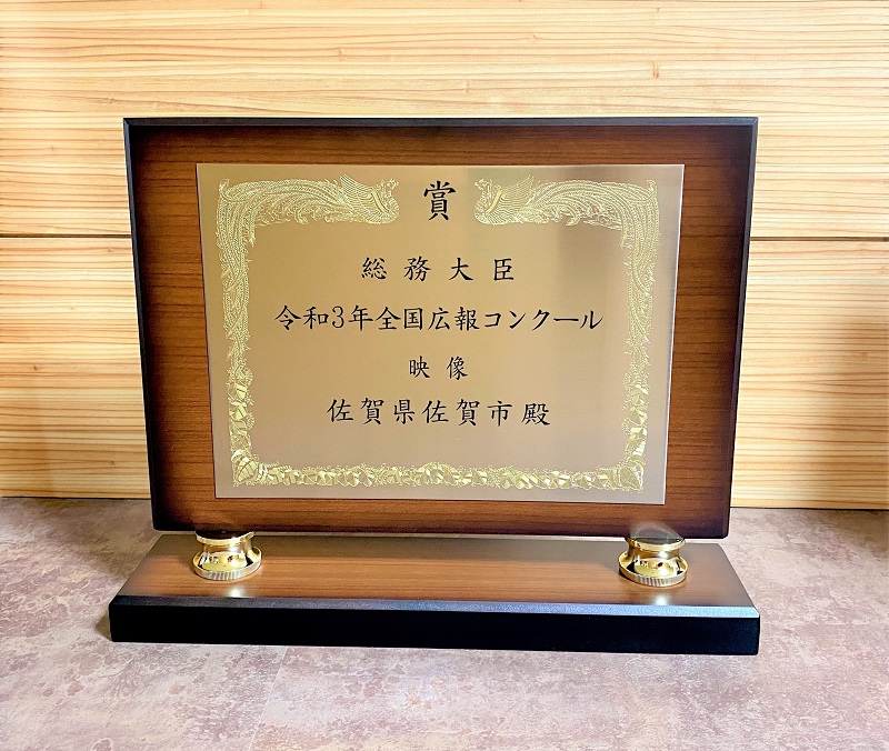 木目調の壁を背景に置かれた、金色のプレートに鳳凰の縁取りが施された表彰盾で、中央には「総務大臣 令和3年全国広報コンクール 映像 佐賀県佐賀市殿」と黒い文字で記されている、栄誉を称える記念の写真