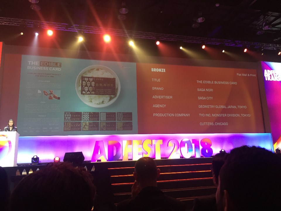 「ADFEST 2018」の華やかな授賞式会場にて、スクリーンに銅賞作品「THE EDIBLE BUSINESS CARD」の詳細情報と、ご飯に乗った海苔の名刺の画像が大きく映し出され、ステージ下部には色鮮やかに光るイベントロゴが配置された授賞式風景写真