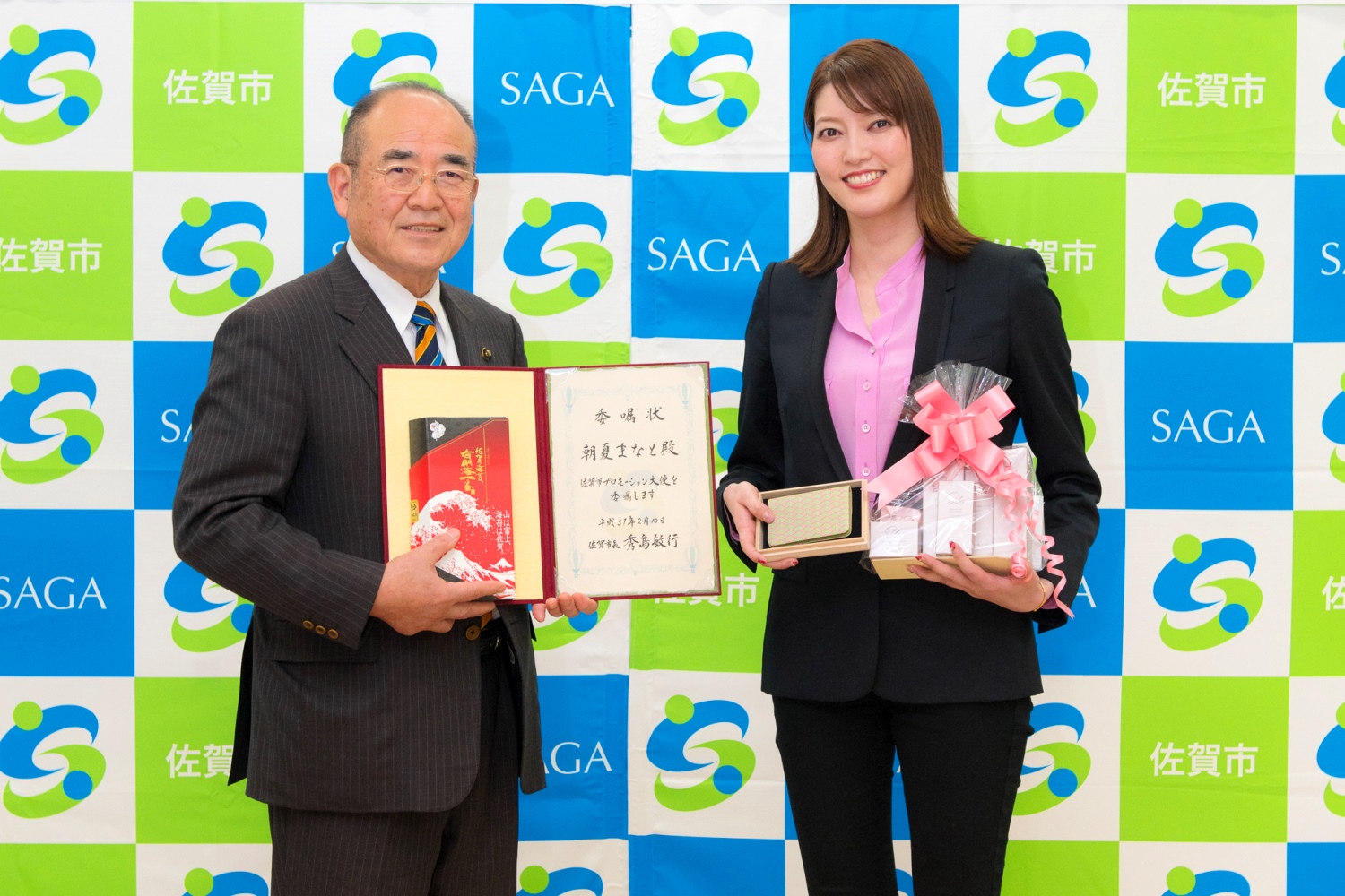 佐賀市のロゴが格子状に並ぶ背景の前で、委嘱状と海苔をもった秀島市長と、Silaのスキンケア&サプリメントセットと名刺入れをもった朝夏まなとさんが並んでいる記念写真