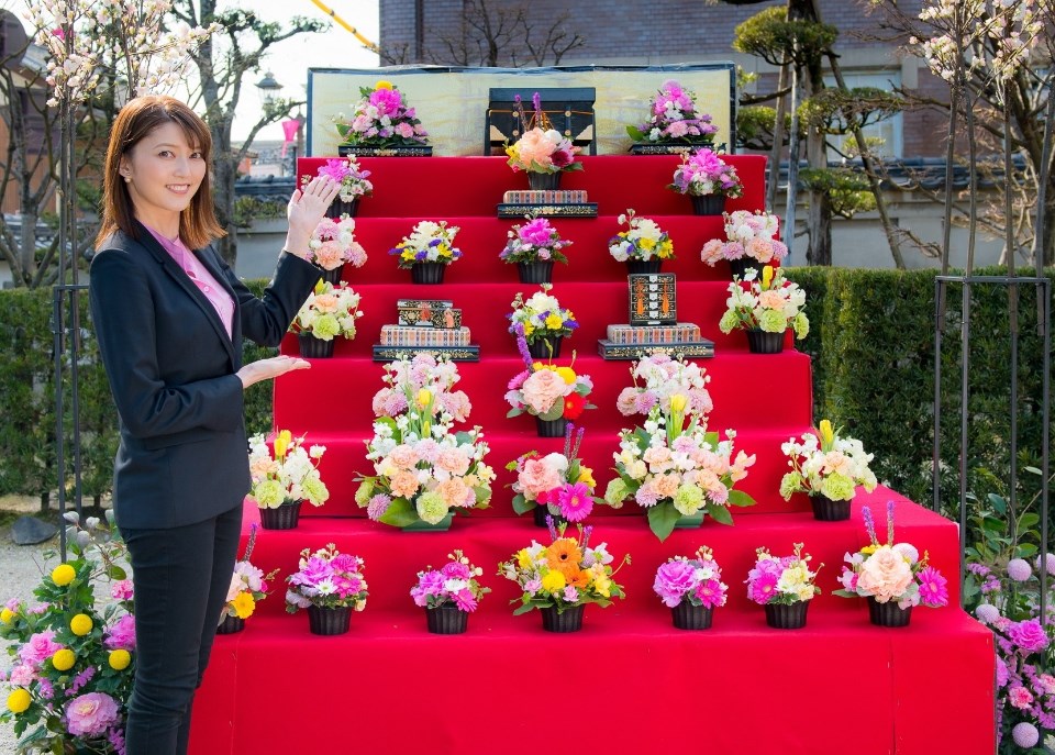 屋外の赤い雛壇に飾られた桃色や黄色の花々の前に、黒いジャケット姿の朝夏まなとさんが立ち、両手を添えるように差し出しながら花雛壇を紹介するように微笑んでいる写真