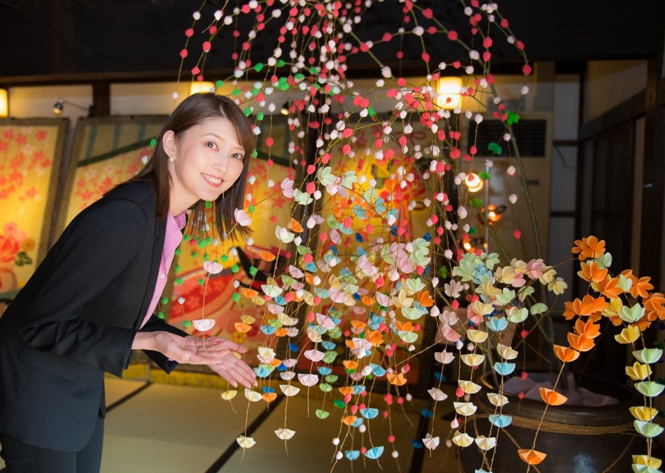 畳が敷かれた和風建築の展示室内で、黒いジャケット姿の朝夏まなとさんが、青地に伝統的な花菱紋様が美しく織り込まれた鍋島緞通を背にして、両手を広げながら晴れやかな笑顔で紹介している写真