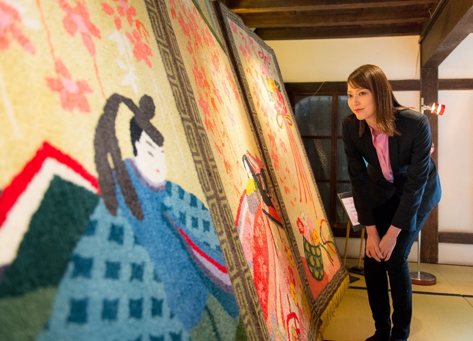 重厚な紋様が施された鍋島緞通が展示されている和室で、朝夏まなとさんが紺色の絨毯の前に立ち、幾何学模様と花々が調和した繊細な織り目を指し示しながら、その風合いを確かめるように微笑んでいる写真