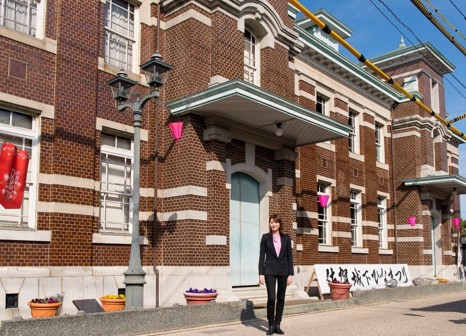 旧古賀銀行の重厚な外観を背に、朝夏まなとさんが建物の魅力を紹介するように、リラックスした自然な立ち姿でカメラを見つめている写真