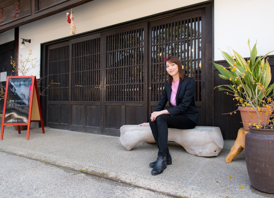 格子状の引き戸が並ぶ歴史的な建物の前で、黒いジャケット姿の朝夏まなとさんが白いベンチに座り、傍らには植物の鉢植えや「和紅茶専門店 紅葉」と書かれた赤い看板が置かれた、歴史ある街並みの雰囲気を感じさせる屋外での全身写真