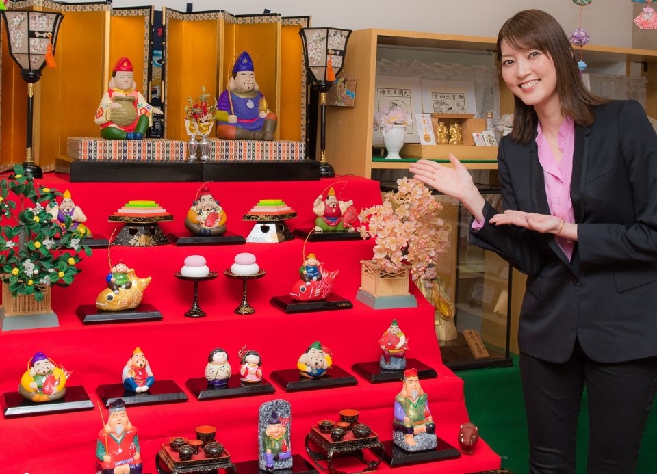 赤い毛氈が敷かれた雛壇のような飾り台に、色鮮やかな張り子の恵比須像や紅白の鏡餅、桜の飾りなどが賑やかに並べられた様子を、黒いジャケット姿の朝夏まなとさんが笑顔で紹介している写真