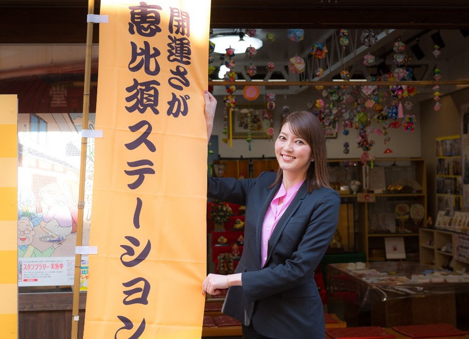 「開運さが恵比須ステーション」と大きく書かれた明るいオレンジ色ののぼり旗の横で、黒いジャケットとピンクのシャツを着用した朝夏まなとさんが、吊るし飾りのある店内を背景に旗を手に持ちながら笑顔を見せている写真
