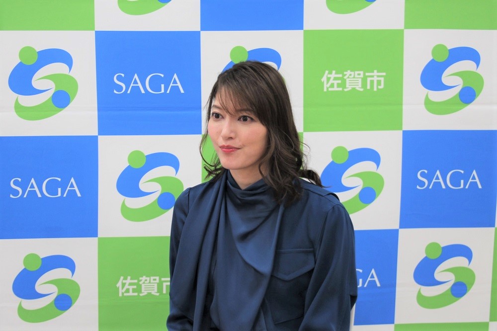 「佐賀市」や「SAGA」の文字、緑と青のロゴマークが交互に並ぶバックパネルの前に、光沢のある紺色のブラウスを身に纏ったセミロングヘアの朝夏まなとさんが立ち、わずかに口角を上げて左側を見つめている上半身の写真