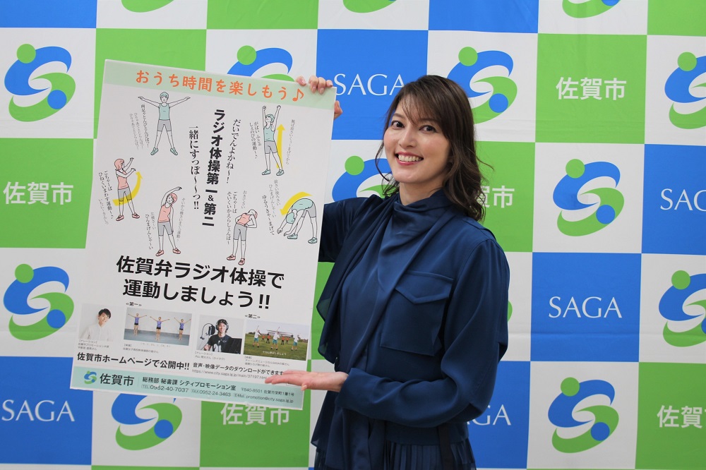 佐賀市のロゴマークが並ぶ背景の前で、紺色のブラウスを着た朝夏まなとさんが「佐賀弁ラジオ体操で運動しましょう!!」と書かれた啓発ポスターを手に持ち、晴れやかな笑顔でこちらに紹介している様子を捉えた宣伝写真