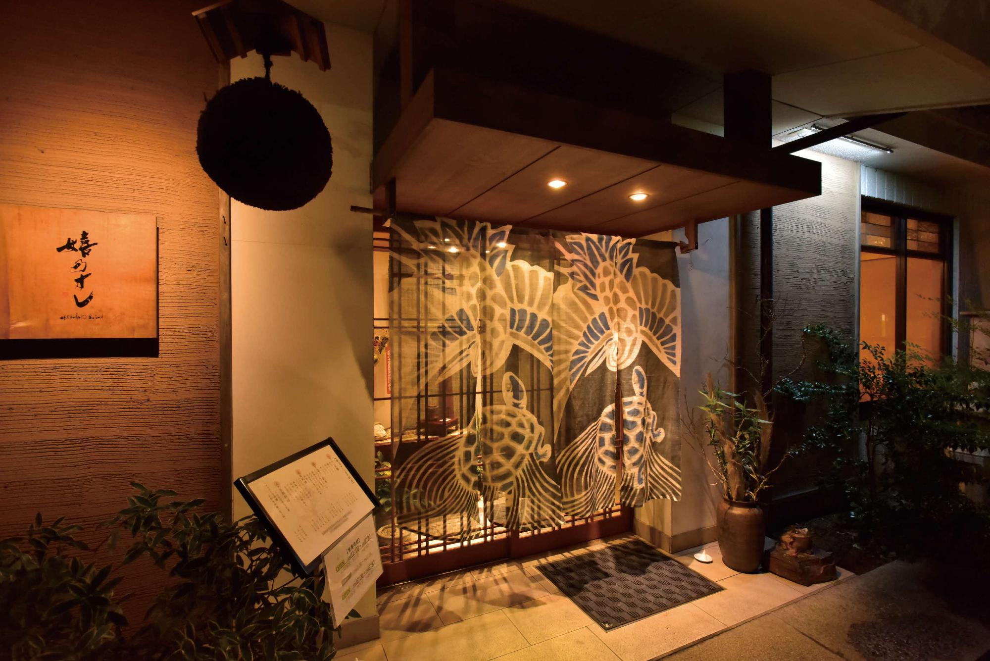 夜の温かな照明に照らされた「嬉乃すし」の店舗入り口で、亀や鶴の縁起の良い文様が描かれた暖簾が下がり、左手には杉玉やメニューボードが置かれた、落ち着いた和の情緒が漂う老舗寿司店の店構えを捉えた写真