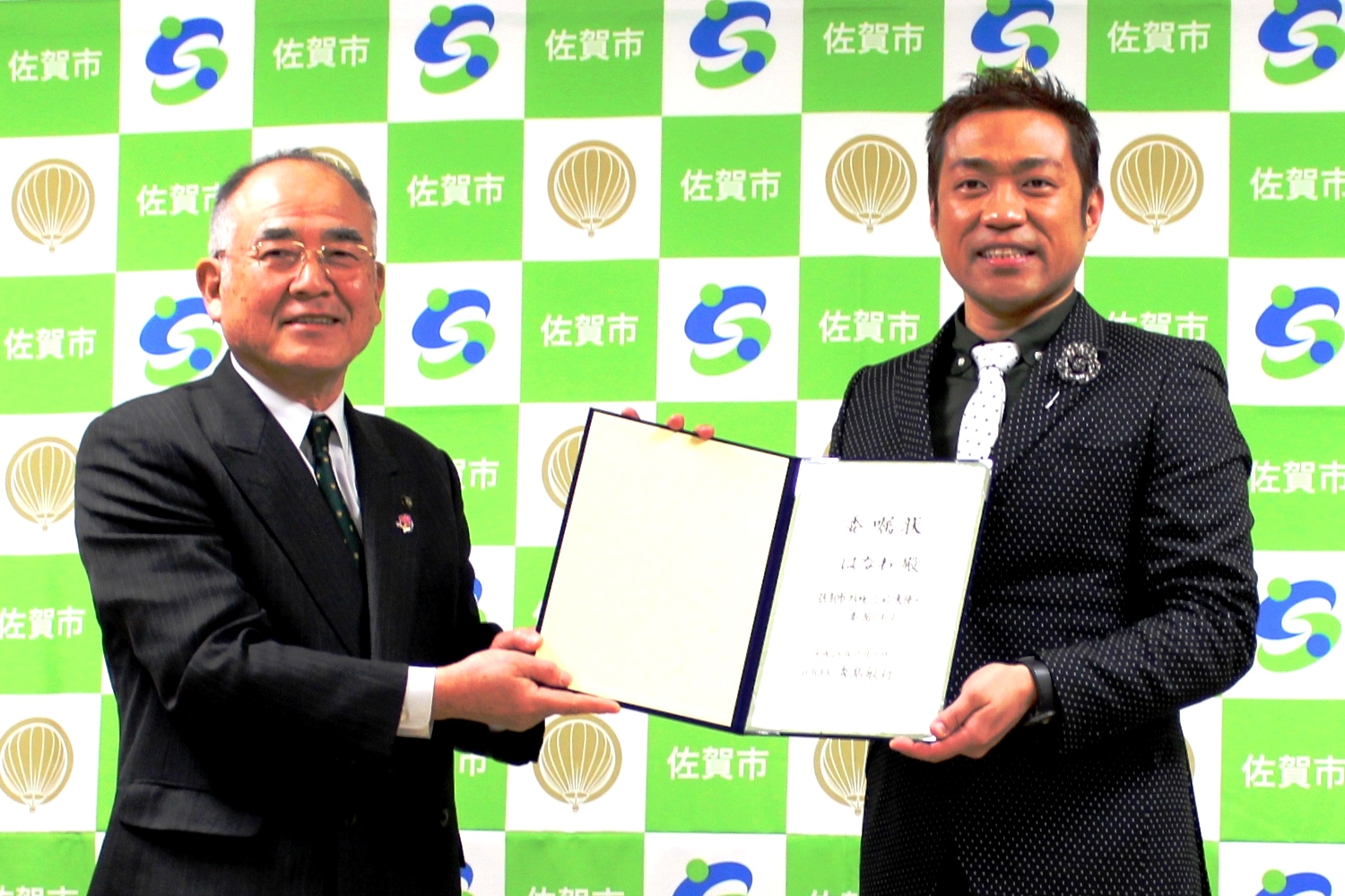 佐賀市のロゴと熱気球のマークが並ぶ背景の前で、スーツ姿の年配の男性と、ドット柄のネクタイに黒いジャケットを着たはなわさんが、二人の間に広げられた委嘱状を手に持ち、並んでいる記念写真