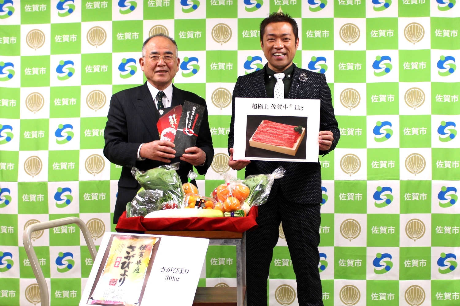 前方に色鮮やかで豊富な野菜果物が整然と並べられ、その後ろにスーツ姿の男性とはなわさんが並んで立ち、肉の写真や特産品を掲げている写真