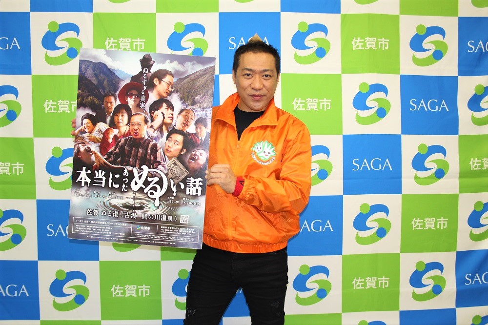 佐賀市のロゴと「SAGA」の文字が並ぶ市松模様のバックパネルを背に、オレンジ色のジャンパーを着た佐賀市プロモーション大使のはなわさんが、多様な人物の顔が並ぶ「本当にあったぬるい話」のキャンペーンポスターを両手で持って掲げている写真