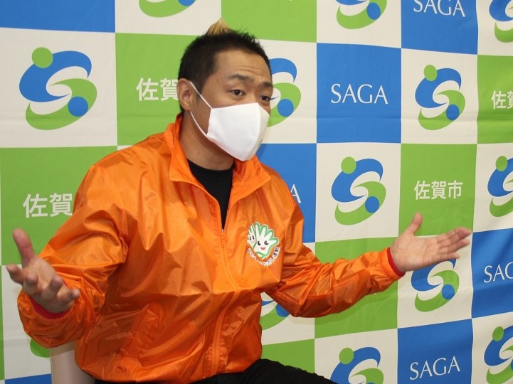 青と緑の市松模様に佐賀市のロゴが並ぶ背景の前で、白いマスクを着用したはなわさんが、胸元にキャラクターのロゴが入った鮮やかなオレンジ色のジャンパーを着て、両手を広げながら熱心に話をしている様子の写真