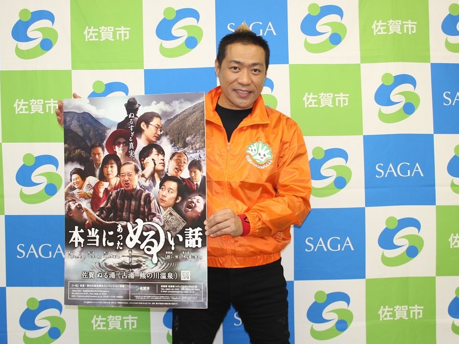 青と緑の市松模様に佐賀市のロゴが並ぶ背景の前で、鮮やかなオレンジ色のジャンパーを着たはなわさんが、多くの人物の顔写真が並ぶ「本当にあったぬるい話」と題された映画ポスター風のパネルを手に持ち微笑む写真