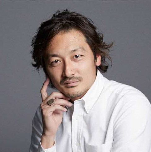 茶髪で緩いウェーブのかかった短髪に髭を蓄え、白いシャツを着た三寺雅人氏が右手の人差し指を顎に添えて思慮深い表情でカメラを見つめている、グレーの背景で撮影されたポートレート写真