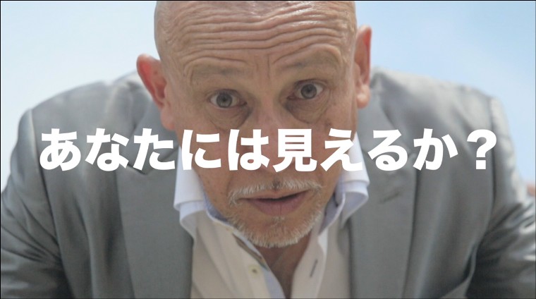 グレーのスーツを着たスキンヘッドの男性が、目を見開きながら真剣な表情でこちらを正面から覗き込んでおり、その顔に重なるように「あなたには見えるか?」という白い大きなテロップが中央に配置された写真