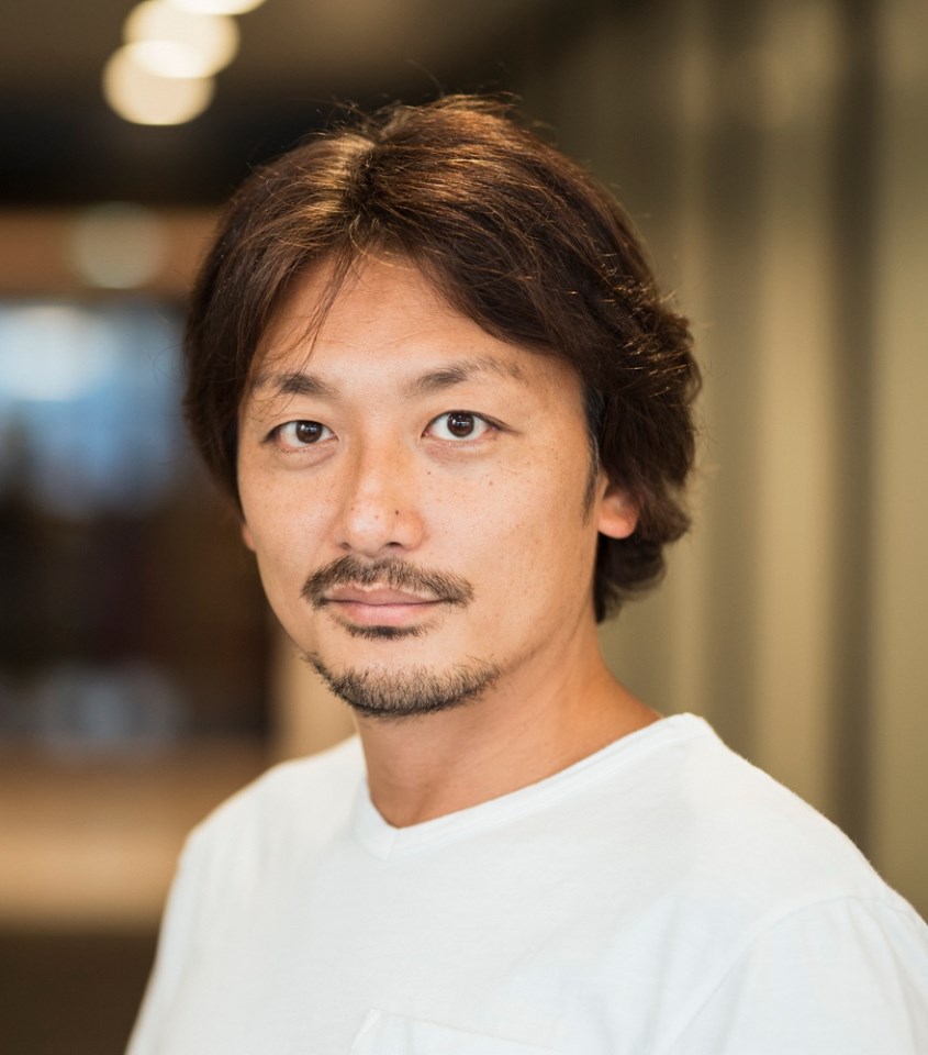 明るいベージュの背景を背に、白いVネックのTシャツを着用した茶髪で髭のある三寺雅人氏が、穏やかな表情で正面を見つめている清潔感のあるポートレート写真