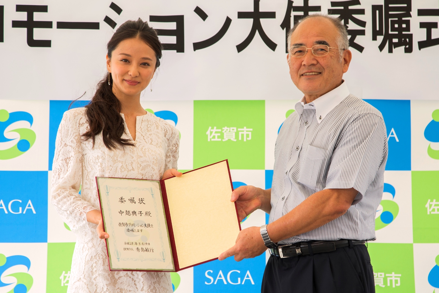 佐賀市のロゴが並ぶ背景の前で、白いレースの服を着た中越典子さんが秀島敏行佐賀市長から「佐賀市プロモーション大使」の委嘱状を受け取り、二人で並んでカメラへ向けて披露する委嘱式の写真