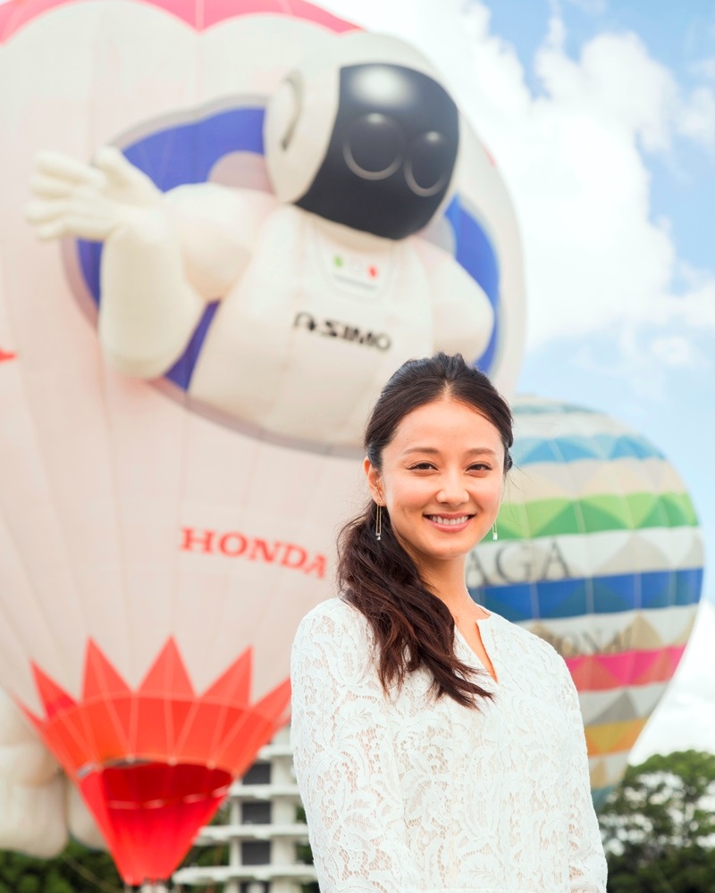 晴天の屋外でHONDAのASIMOを模した巨大な熱気球を背景に、白いレースの服を着た中越典子さんが微笑みを浮かべて佇むポートレート写真