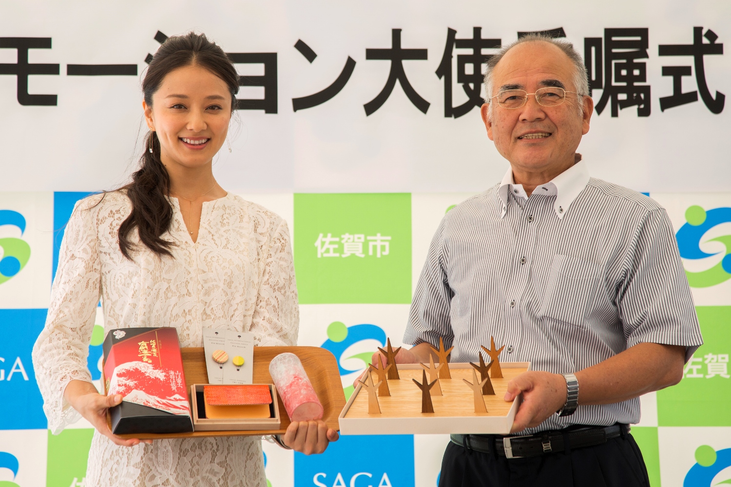 佐賀市のロゴが並ぶ背景の前で、白いレースの服を着た中越典子さんと秀島敏行市長が、佐賀海苔や佐賀錦の名刺入れ、諸富家具の木製グッズなど大使就任の記念品を手に並んで微笑む、佐賀市プロモーション大使委嘱式の写真