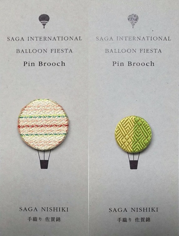 「SAGA INTERNATIONAL BALLOON FIESTA(佐賀インターナショナルバルーンフェスタ)」と印字された2枚のグレーの台紙に、左側はピンクや赤の縞模様、右側は黄緑色の幾何学模様のデザインの伝統的な佐賀錦の織地で作られた円形のピンブローチが、熱気球の球体部分に見立ててそれぞれ1つずつ固定されている写真