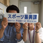 「スポーツ振興課 Sports Promotion Division」と青い文字で書かれた白い看板を、男女二人が顔を隠すように正面に掲げて持っている写真