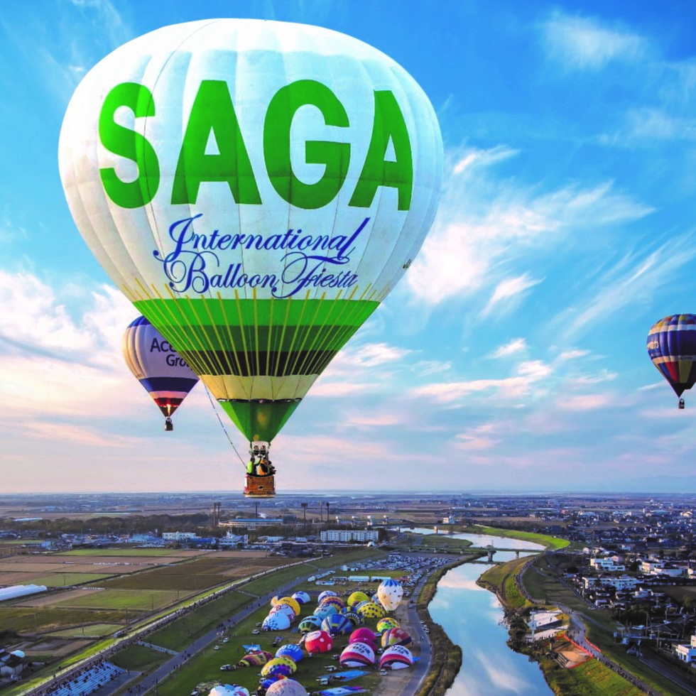 青空が広がる佐賀平野の上空を「SAGA International Balloon Fiesta」と書かれた白と緑の気球が飛行しており、眼下には川や田畑、そして離陸を待つ色とりどりの多数の気球が並ぶ大会風景を捉えた写真