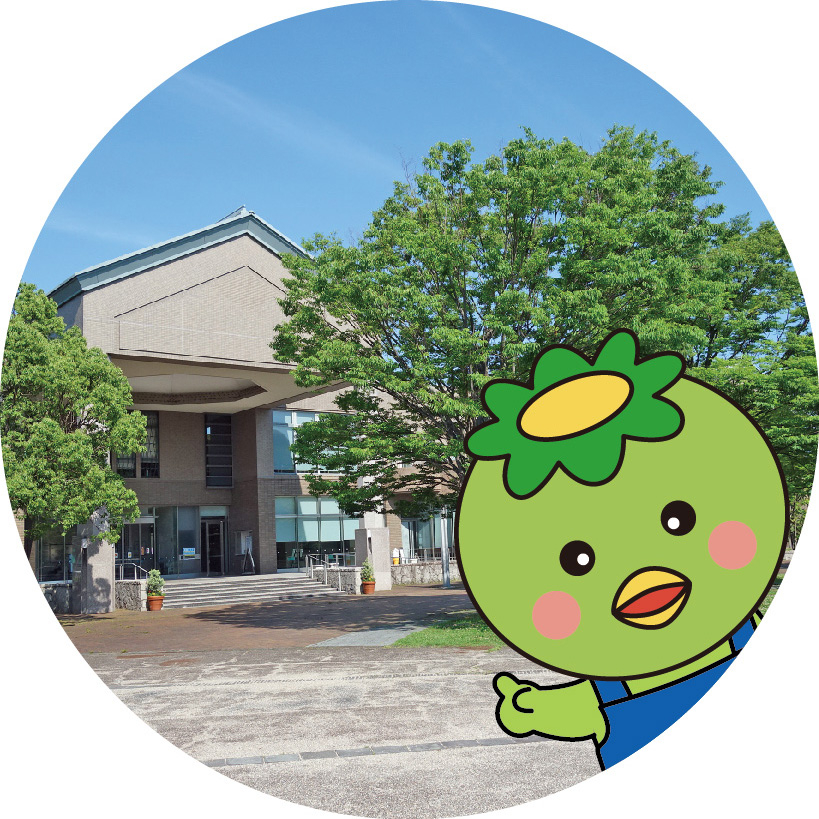ベージュ色の大きな公共施設と緑豊かな木々を背景に、手前右側から緑色の体に黄色い花のような帽子を被ったカッパのキャラクターがひょっこりと顔を出して施設を指差している合成写真