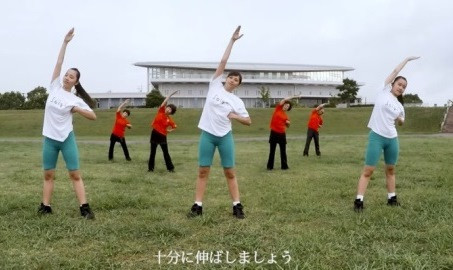 芝生が広がる屋外で、白いTシャツと緑のハーフパンツを着用した三人の若い女性を中心に、後方の赤いTシャツを着た年配の女性たちと一斉に片腕を高く上げ、体を横に曲げる運動を行う様子を捉えた実演写真