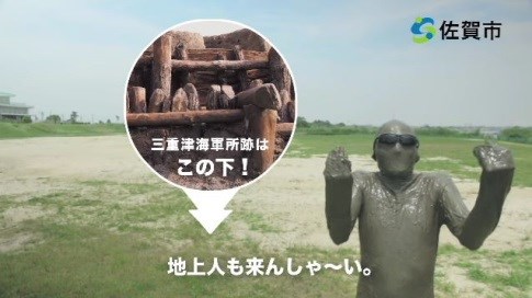 河川敷で全身泥まみれの人物がポーズをとり、画面には三重津海軍所跡の遺構写真と「この下!」という指示、さらに「地上人も来んしゃ~い。」という字幕と佐賀市のロゴが表示された、歴史遺産をPRする切り出し写真