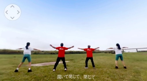 広々とした緑の草地で、赤いTシャツの男性二名と白いTシャツの女性二名が背を向け、青空の下で両腕を水平に大きく広げ足を開く運動を行っている様子を捉え、下部に「開いて閉じて、閉じて」と字幕を添えた実演写真