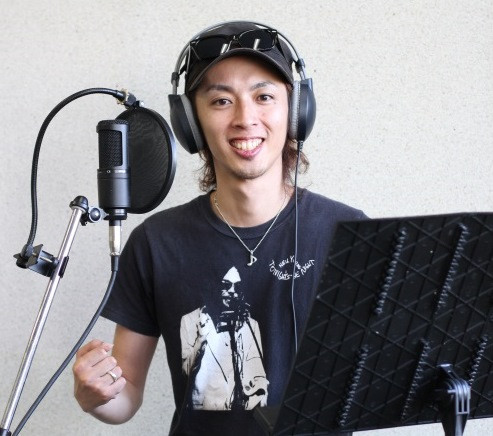 録音スタジオでヘッドフォンとサングラス付きの帽子を着用した男性が、マイクの前で笑顔を見せ、右手に握りこぶしを作ってやる気を表現している、佐賀弁ラジオ体操第二のナレーションを担当する内山敬太さんの紹介写真
