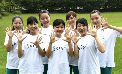 芝生が広がる屋外で、「Soigi」と書かれたお揃いの白いTシャツを着た若い女性7人が、カメラに向かって両手を広げ、弾けるような笑顔でポーズをとっている様子を捉えた、佐賀弁ラジオ体操の実演グループの集合写真