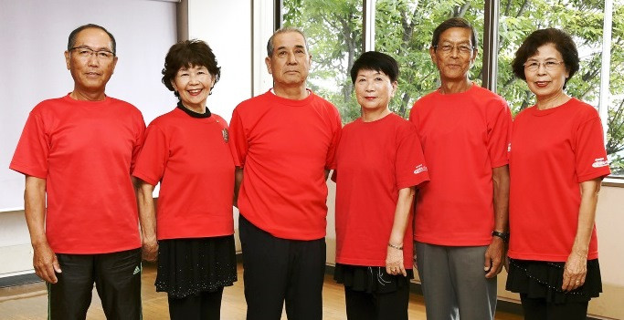 赤いTシャツを着た年配の男女六名が屋内の板張りの床に並んで立ち、窓から見える木々の緑を背景に、穏やかな笑顔や真剣な表情でカメラを見つめている、佐賀弁ラジオ体操の実演グループ「健康ひろげ隊」の集合写真