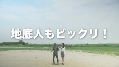 広々とした河川敷の砂地に、全身が泥で覆われた人物と白い服にチェック柄のスカートを履いた女性が並んで立っており、画面中央には「地底人もビックリ!」という白い大きなテロップが重なっている写真