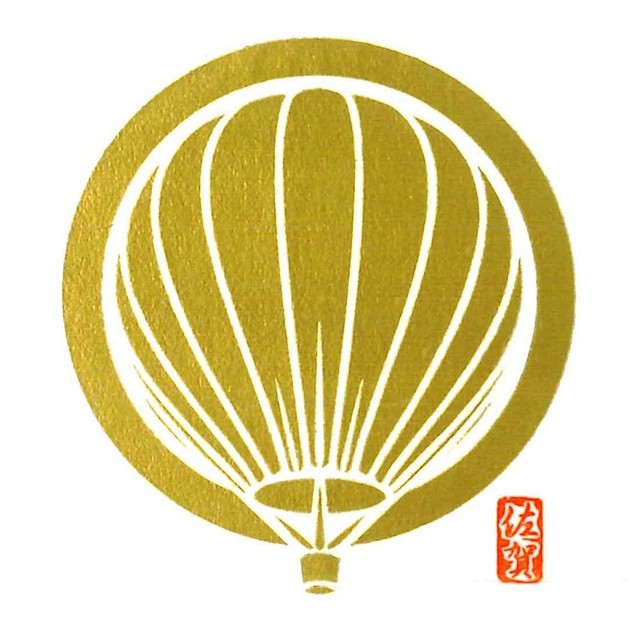 金色の円形の中に、上昇する熱気球の輪郭が白抜きの線画で繊細に描かれ、右下には赤地に白抜きで佐賀と記された小さな落款印が配置されたイラスト