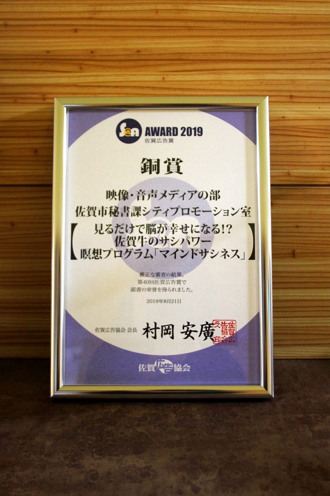 木目調の壁を背景に、映像・音声メディアの部にて佐賀市秘書課シティプロモーション室の「マインドサシネス」が受賞したことを示す、シルバーの額縁に収められた「第40回佐賀広告賞」の銅賞の賞状が立てかけられた写真