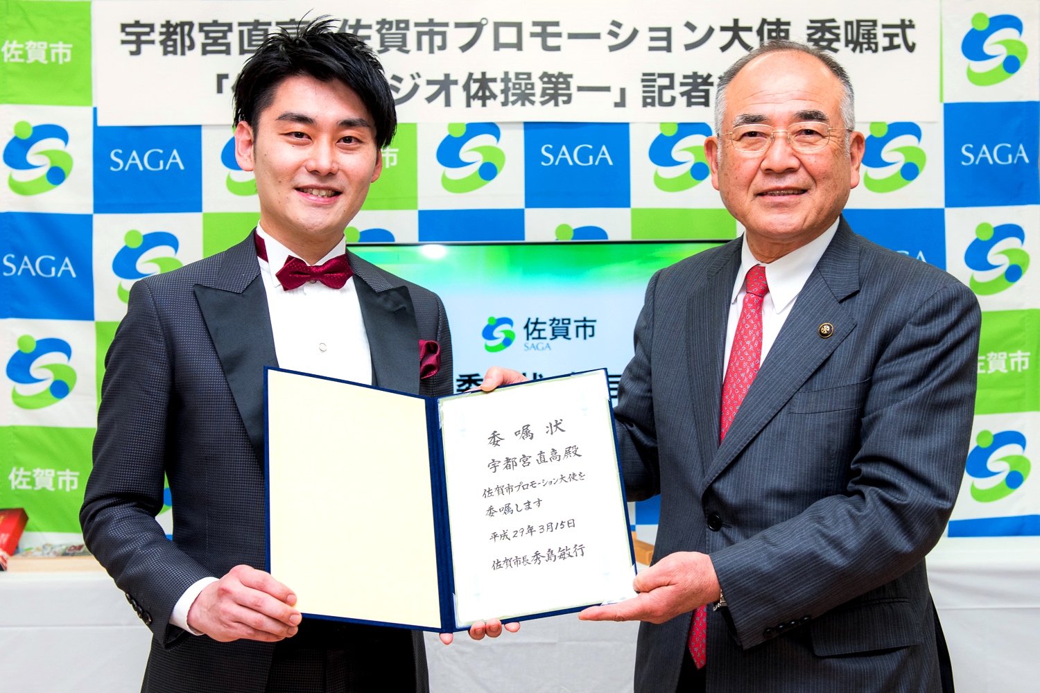 佐賀市プロモーション大使委嘱式にて、宇都宮直高氏へ委嘱状を授与する秀島敏行佐賀市長が、佐賀市のロゴが並ぶ背景の前で、二人で開いた委嘱状を持ちながら笑顔で並んで立っている、公式行事での記念写真