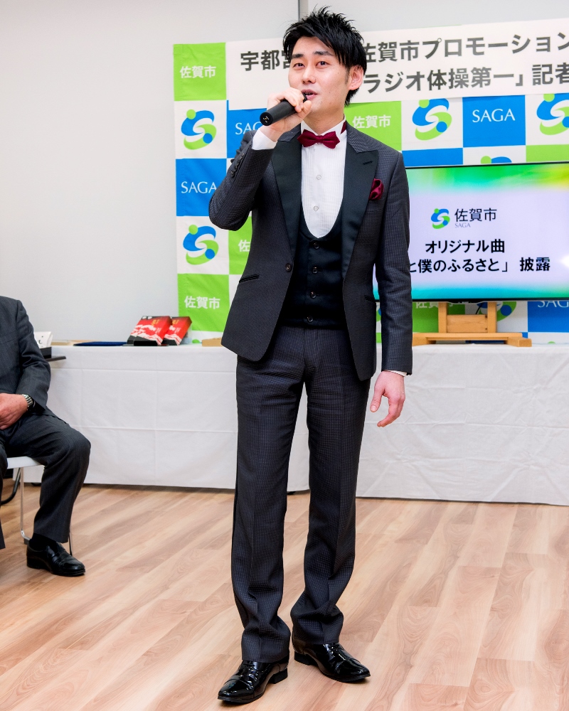 佐賀市プロモーション大使の宇都宮直高氏が、佐賀市のロゴが並ぶ背景の前でマイクを手に取り、タキシード姿でオリジナル曲を披露している様子の写真
