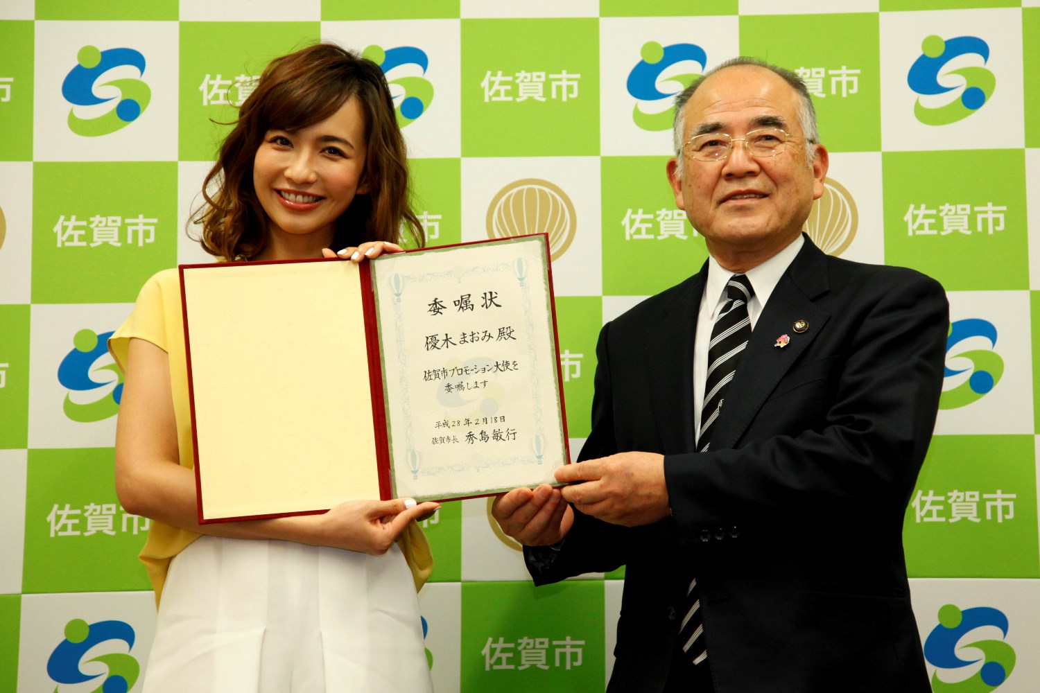 佐賀市のロゴと市名が並ぶ市松模様のバックパネルの前で、佐賀市プロモーション大使の委嘱状を手に持つ優木まおみさんと、その隣で微笑みながら委嘱状を支えるスーツ姿の秀島市長が並んで立っている記念写真