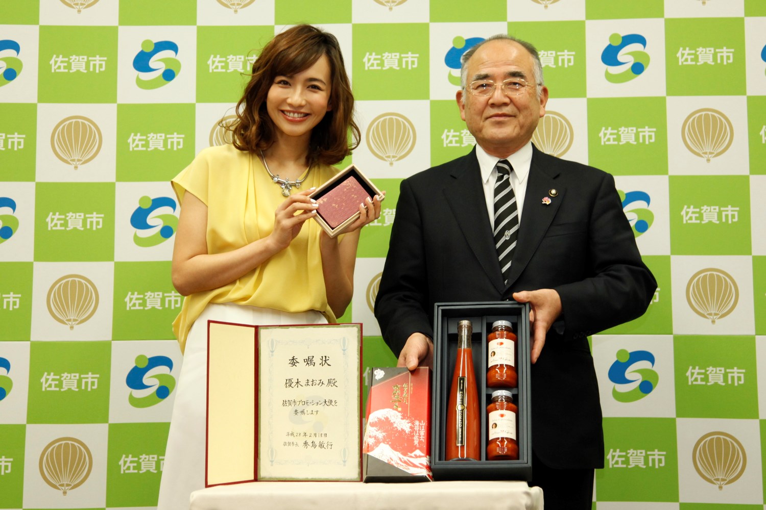 佐賀市のロゴと市名が並ぶ市松模様のバックパネルの前で、優木まおみさんが記念品を手に持ってスーツ姿の市長の隣で微笑み、手前には委嘱状や特産品のボトルセットが並べられた、佐賀市プロモーション大使の就任を記念する写真
