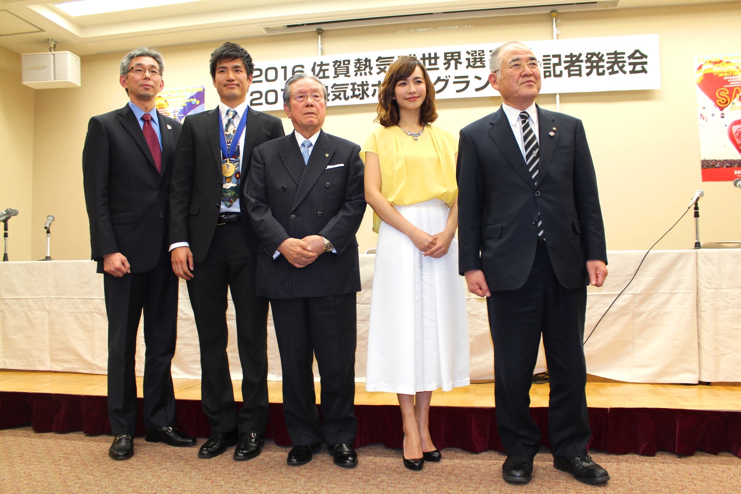 「2016佐賀熱気球世界選手権記者発表会」の看板を背に、優木まおみさんとスーツ姿の秀島市長や世界チャンピオンの藤田雄大さん、関係者の男性2人と横一列に並び、これから始まる大会への期待を感じさせる表情で正面を見据えて立っている、イベント会場での集合写真